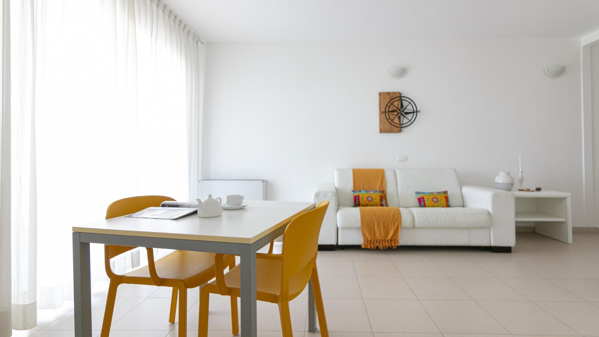 Un salon moderne avec un canapé blanc, des fauteuils jaunes, une table blanche et une décoration murale représentant une boussole.