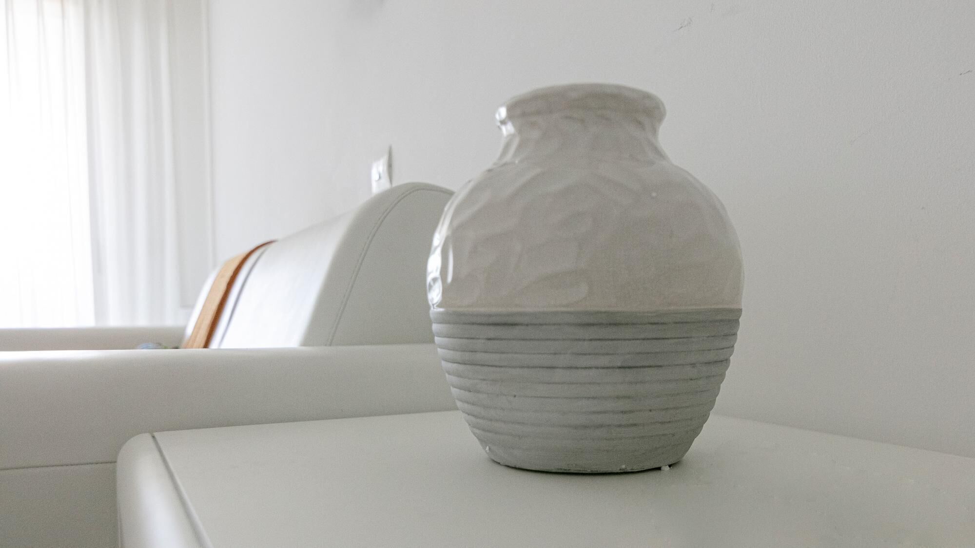 A white ceramic vase on a white table in a room with a white wall and a window with blinds.