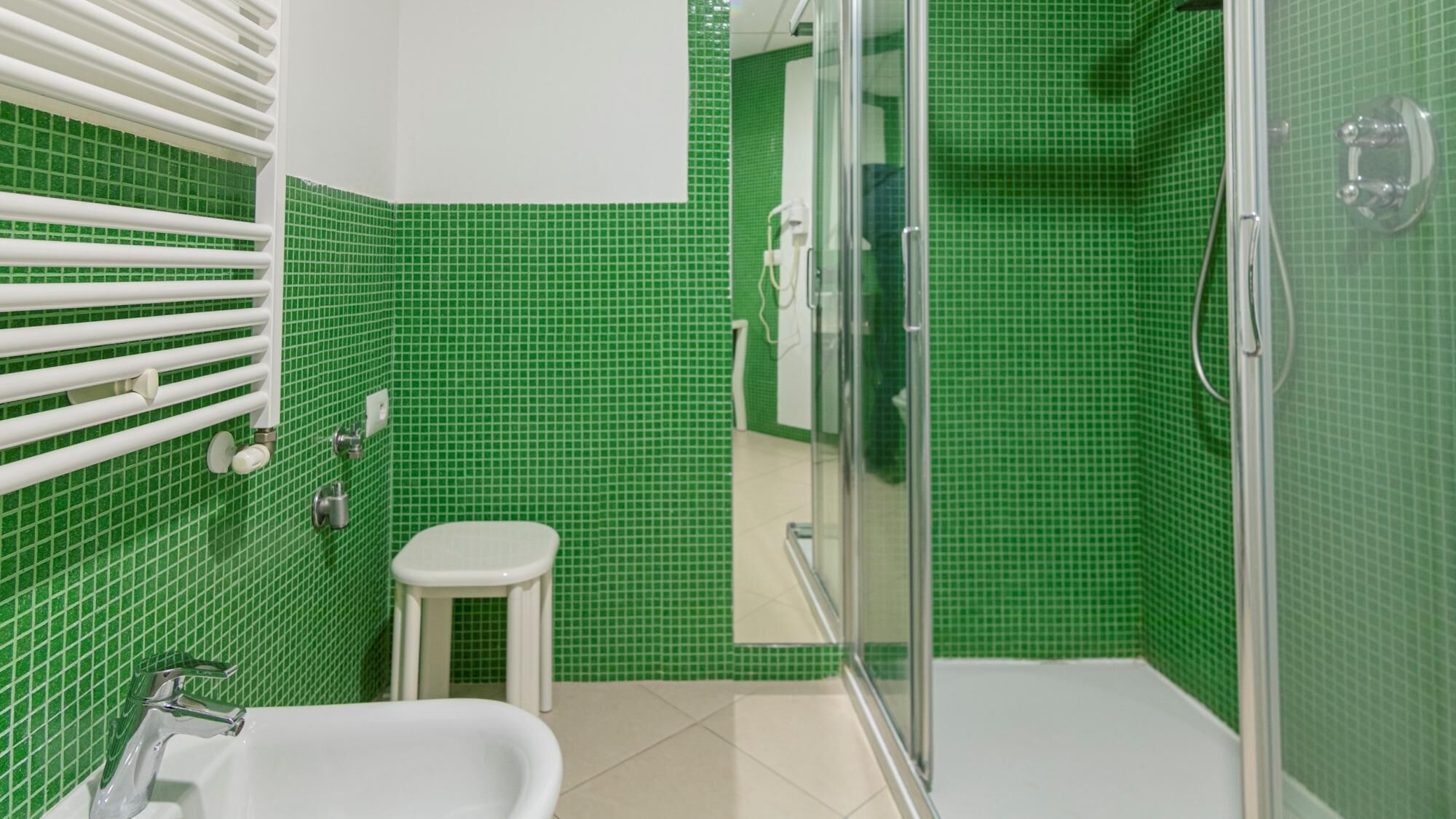 A bathroom with green tiled walls, a white bathtub, a small white stool, and a glass-enclosed shower area.