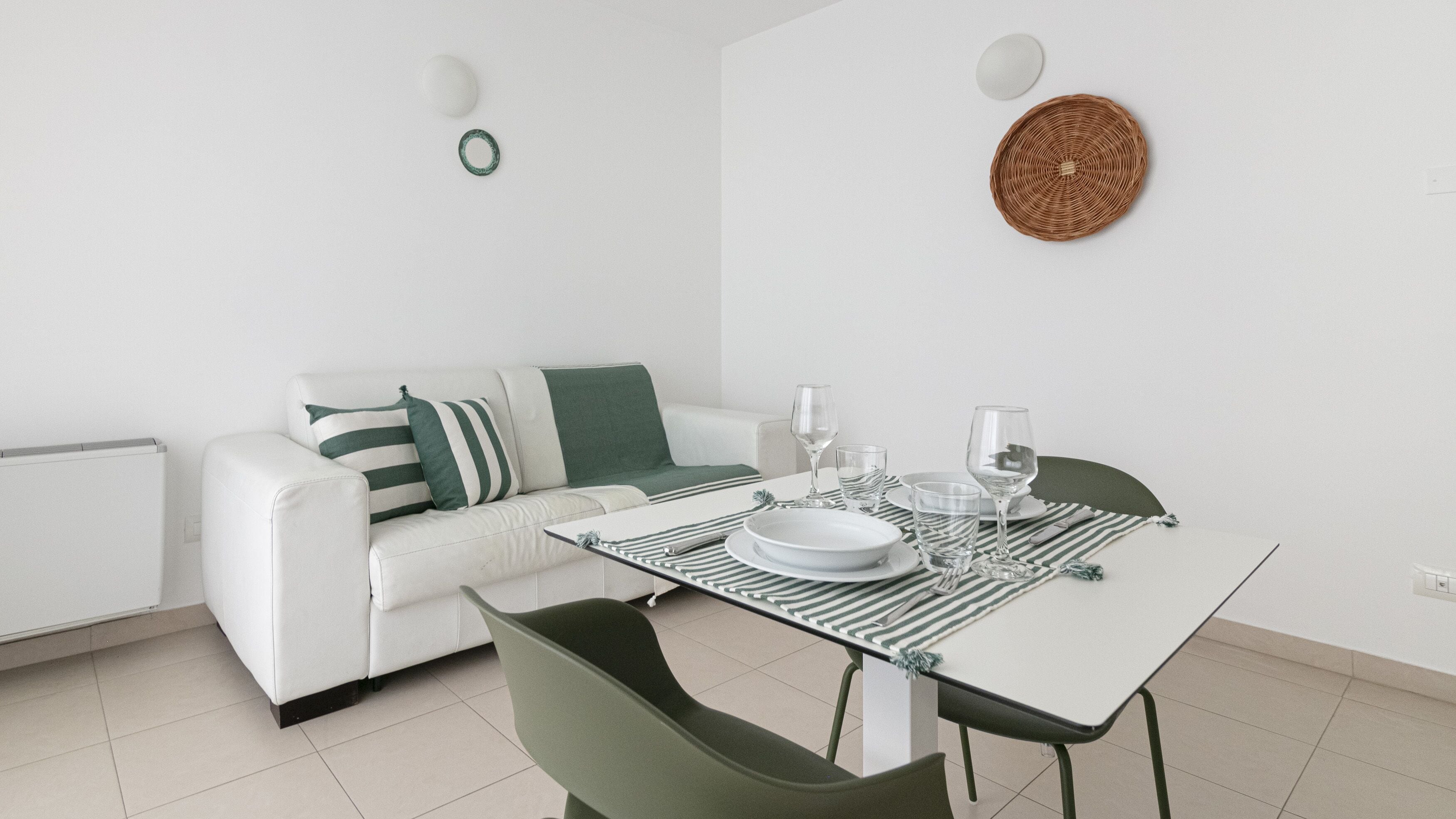 A modern, minimalist living room with a white sofa, green and white striped pillows, a dining table with green chairs, and a woven wall hanging.