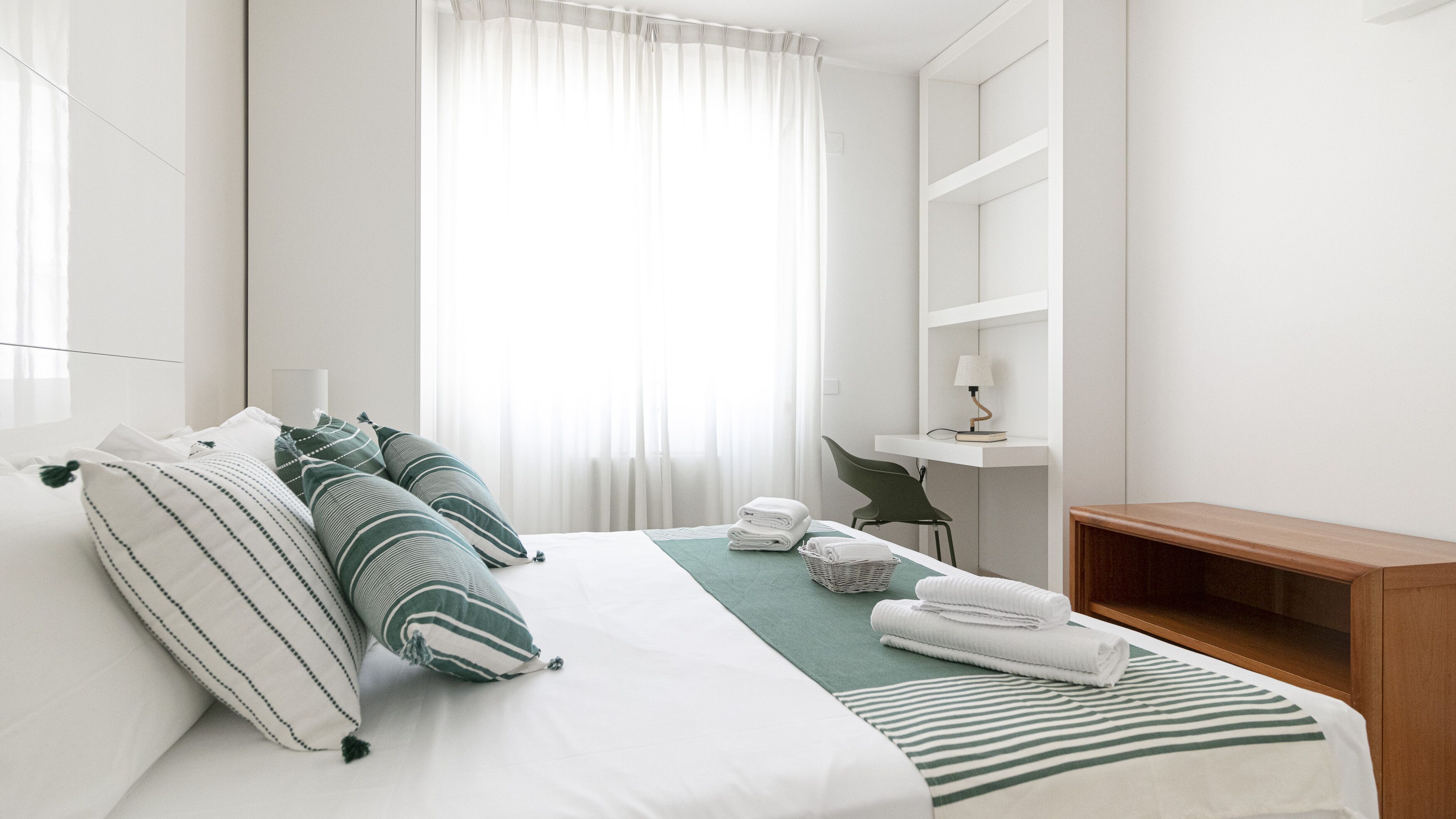 A hotel room with a neatly made bed, a bedside table with a lamp, a chair, and a wooden bench.