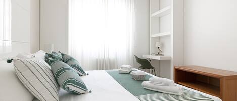 A hotel room with a neatly made bed, a bedside table with a lamp, a chair, and a wooden bench.