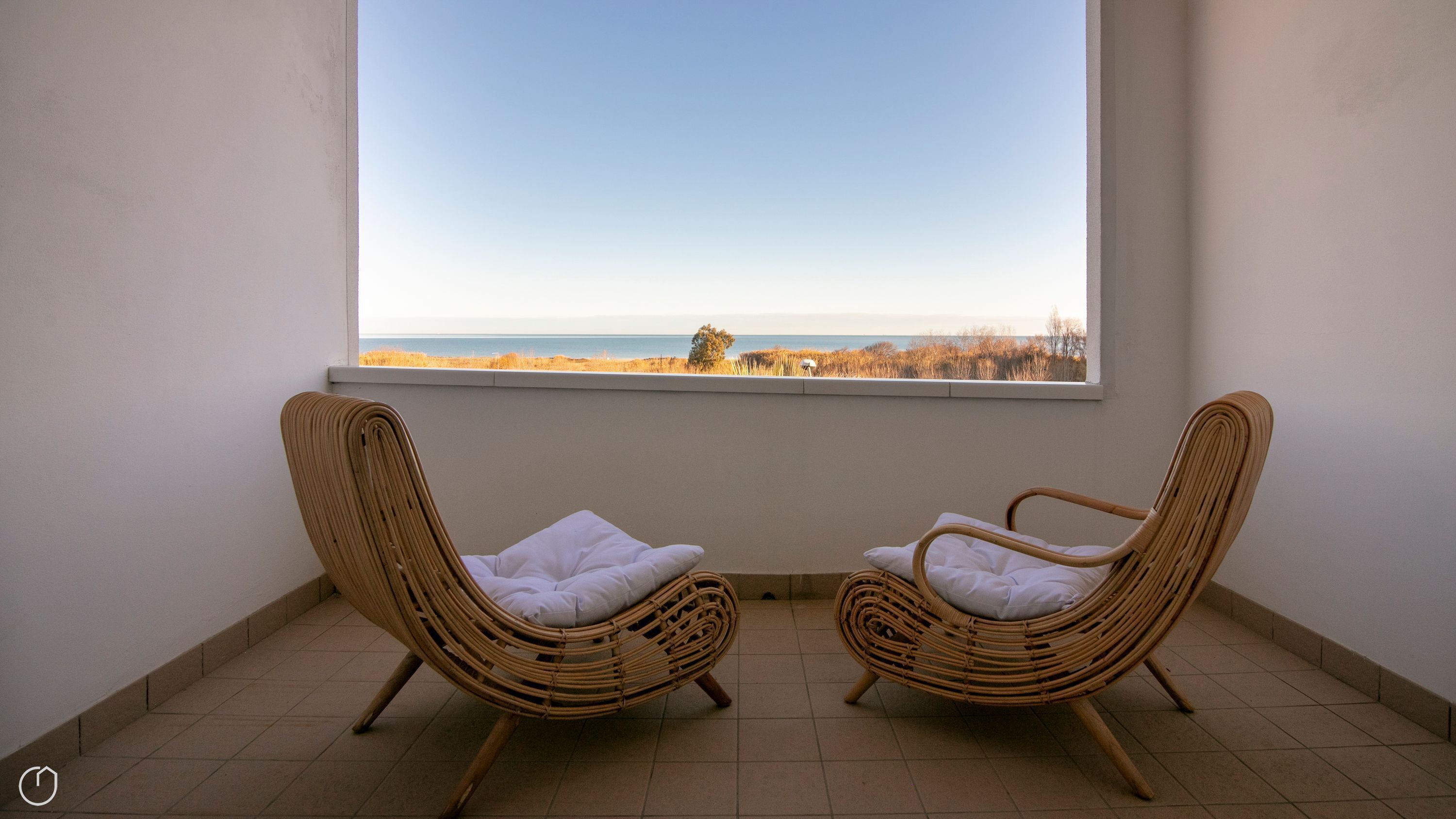 Two wicker chairs with white cushions facing a large window overlooking a beach.
