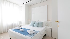 Une chambre moderne avec un grand lit, du linge de lit blanc et une touche de couleur bleue. On y trouve une table de chevet avec une lampe et une petite table en bois.