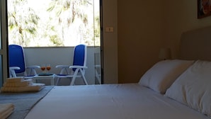 A hotel room with a bed, two blue chairs, a small table with drinks, and a view of palm trees through the window. - Le Barchette 8 - Primo Piano (Lecce)