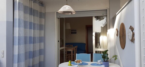 A room with a dining table set for two, a blue chair, a small potted plant, and a hanging light fixture.