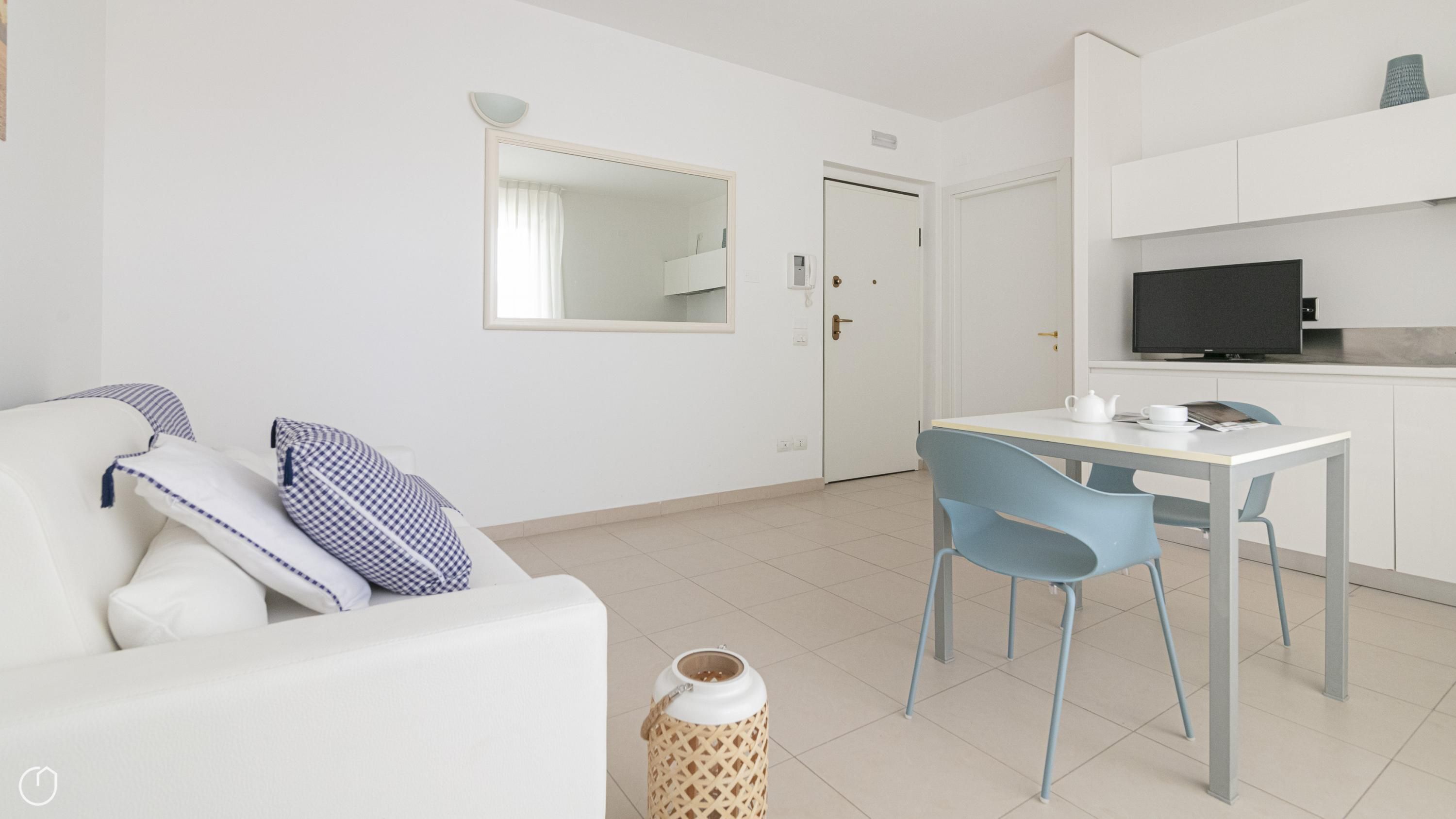 Ein modernes, minimalistisches Wohnzimmer mit einem weißen Sofa, blauen Sesseln, einem kleinen Tisch mit einem Teekessel und einem Fernseher auf einem weißen Schrank.