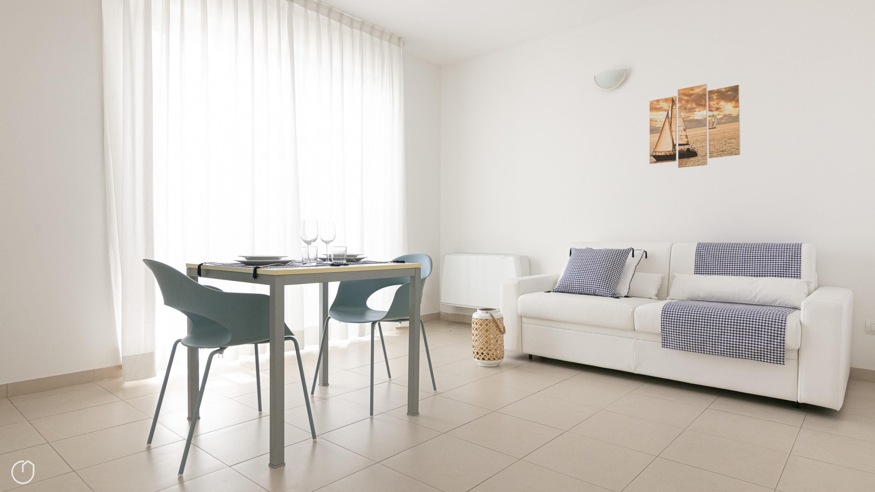 A modern living room with a white sofa, two chairs, a dining table, and a large window with sheer curtains.