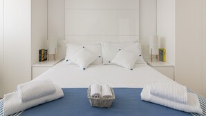 Un lit parfaitement fait, avec du linge de lit blanc et des touches de bleu, un panier rempli de serviettes pliées, et une chambre au design épuré et minimaliste.