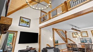 A spacious living area with a high ceiling, wooden beams, a staircase, and a dining area with multiple chairs.