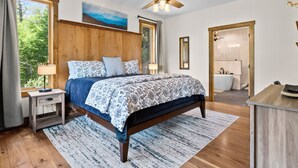 A bedroom with a wooden headboard, a bed with patterned bedding, a nightstand with a lamp, a ceiling fan, and a view of the outdoors through a window. - New Home on South Toe River, Pet Friendly (Burnsville)