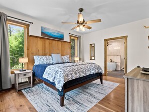 A bedroom with a wooden headboard, a bed with patterned bedding, a nightstand with a lamp, a ceiling fan, and a view of the outdoors through a window.