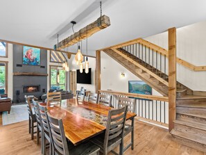 Uma sala de estar ampla, com uma grande mesa de jantar de madeira, assentos confortáveis, uma lareira e uma escada.