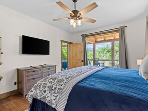 Un dormitorio con cama, cómoda, televisor y balcón con vista a árboles.