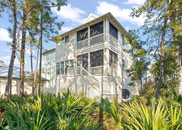 A two-story house with a balcony and a covered porch, surrounded by tall pine trees.