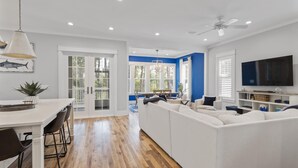 A spacious living room with a white sofa, a dining area with a table and chairs, and a TV mounted on the wall. - Family Beach House w/ LSV and Porches (Santa Rosa Beach)