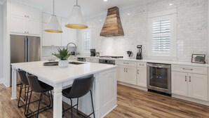 A modern kitchen with white cabinetry, stainless steel appliances, and a central island with bar stools. - Family Beach House w/ LSV and Porches (Santa Rosa Beach)