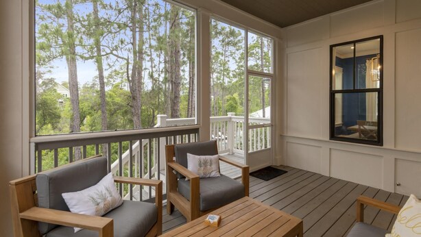 A covered porch with wooden furniture, large windows offering a view of tall trees, and a ceiling fan. - Family Beach House w/ LSV and Porches (Santa Rosa Beach)