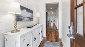 A hallway with white paneled walls, a wooden floor, a white console table with two lamps, and a painting of a seascape. - Family Beach House w/ LSV and Porches (Santa Rosa Beach)