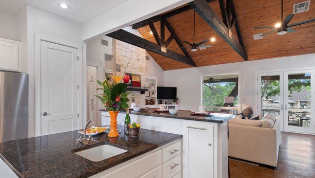 Una cocina moderna con encimera de granito, armarios blancos y horno empotrado. Hay un comedor con sofá y vistas al exterior.