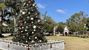 A large Christmas tree decorated with ornaments, surrounded by a white picket fence in a park setting.