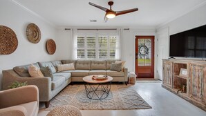 A modern living room with a sectional sofa, a ceiling fan, a large window with blinds, and a television on a wooden cabinet.