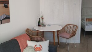 A cozy room with a grey sofa, wicker chairs, a white round table with a bottle and glasses, and a bed with a white headboard. - Luxury apartment for beach/walking  3 minute walk to the beautiful sandy beach. (Egmond aan Zee)