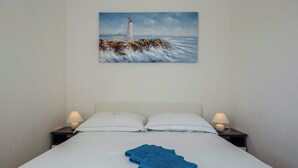 Een net opgemaakt bed met witte lakens en twee kussens, een blauwe handdoek op het bed, en een schilderij van een vuurtoren aan de muur.
