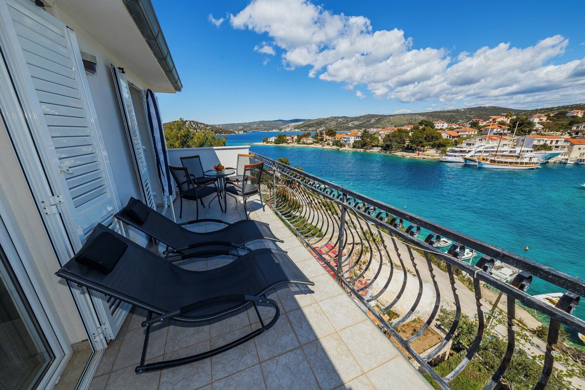 A balcony with two lounge chairs, a small table, and a view of a coastal town and sea.