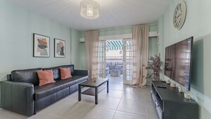 Un salon avec un canapé en cuir noir, une table basse, un téléviseur posé sur un meuble noir et une grande fenêtre avec des rideaux.