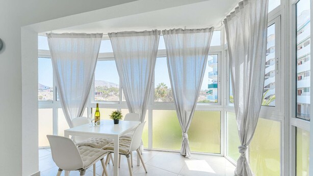 A dining area with a table and chairs, large windows with sheer curtains, and a view of buildings and greenery outside. - Rising Sun Apartment in complex Las Terrazas (Adeje)