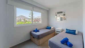 A bedroom with two beds, a small bedside table, a nightstand, and a window with a view of hills and greenery. - Rising Sun Apartment in complex Las Terrazas (Adeje)