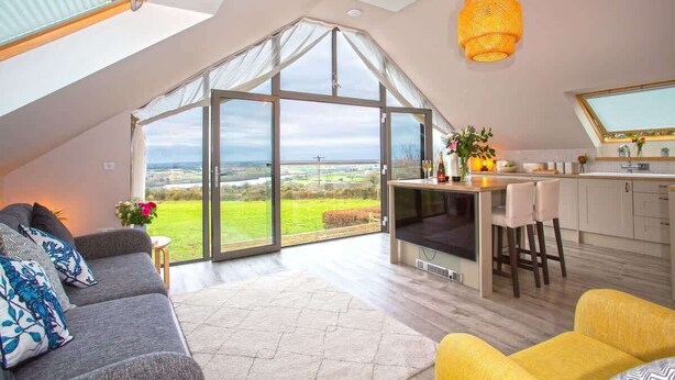 A modern loft with a large glass door leading to a scenic outdoor view, a cozy seating area, and a kitchen with a bar. - The Neyth (Mabe Burnthouse)