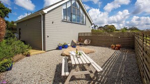 A single-story house with a sloped roof, a large window, and a gravel patio with a wooden table and chairs. - The Neyth (Mabe Burnthouse)