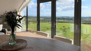 A dining area with a table and chairs, a vase with flowers, and a view of the countryside through large windows. - The Neyth (Mabe Burnthouse)