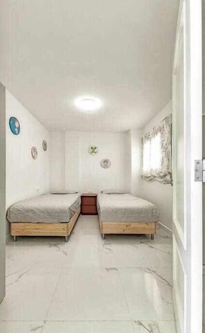 A small, clean room with two single beds, a small table, and a window with curtains. - Moderno apartamento en el Parque Royal I (Adeje)