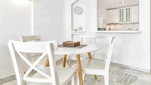 A dining area with white chairs and a round table, a wall-mounted clock, and a kitchen visible through a window. - Apartment in complex Altamira with direct sea view (Costa Adeje)