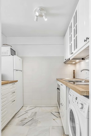 A modern kitchen with white cabinetry, a built-in oven, a microwave, a washing machine, and a wooden countertop. - Apartment in complex Altamira with direct sea view (Costa Adeje)