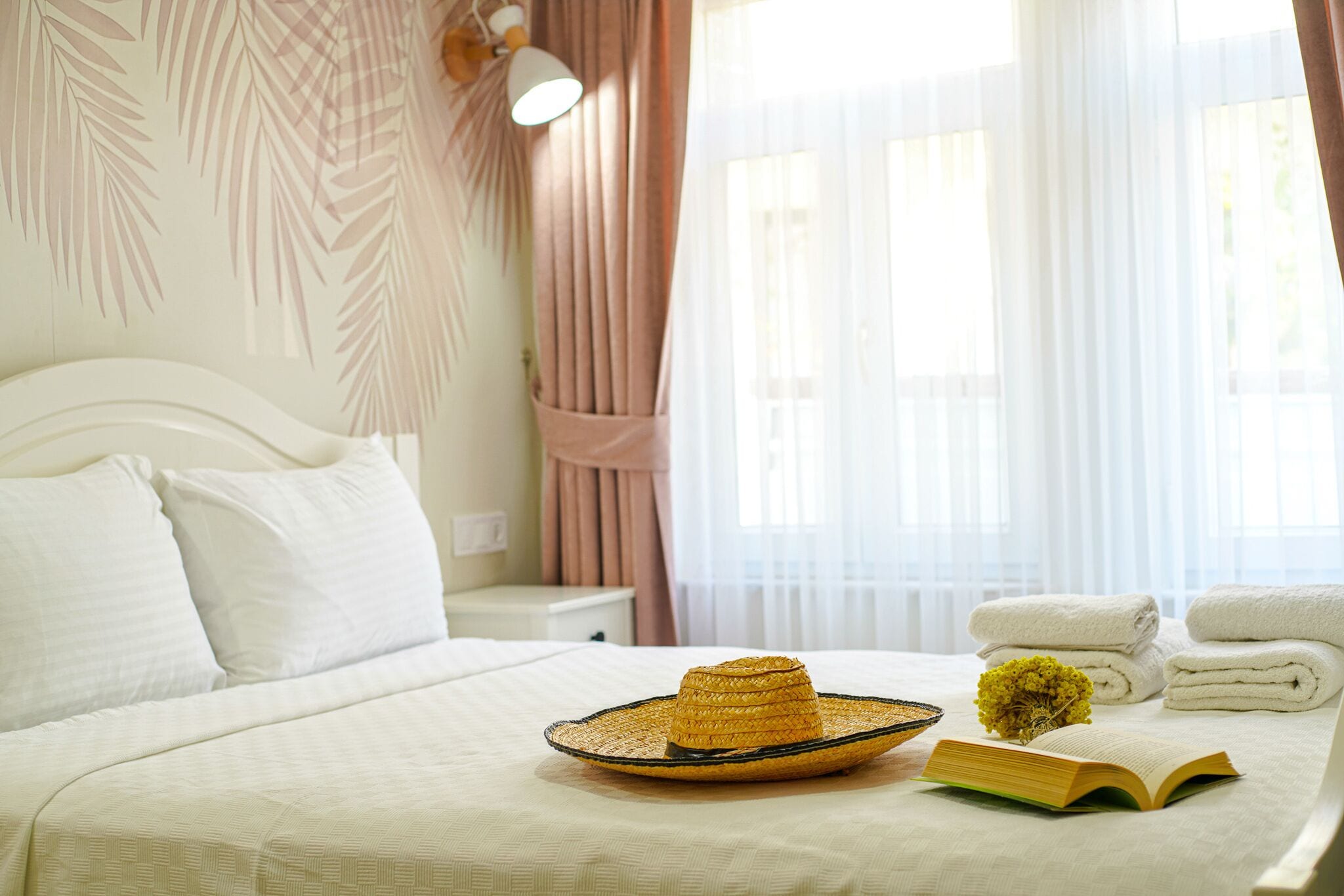 Una cama bien hecha con sábanas blancas, un sombrero de paja, un libro y toallas en una habitación de hotel.