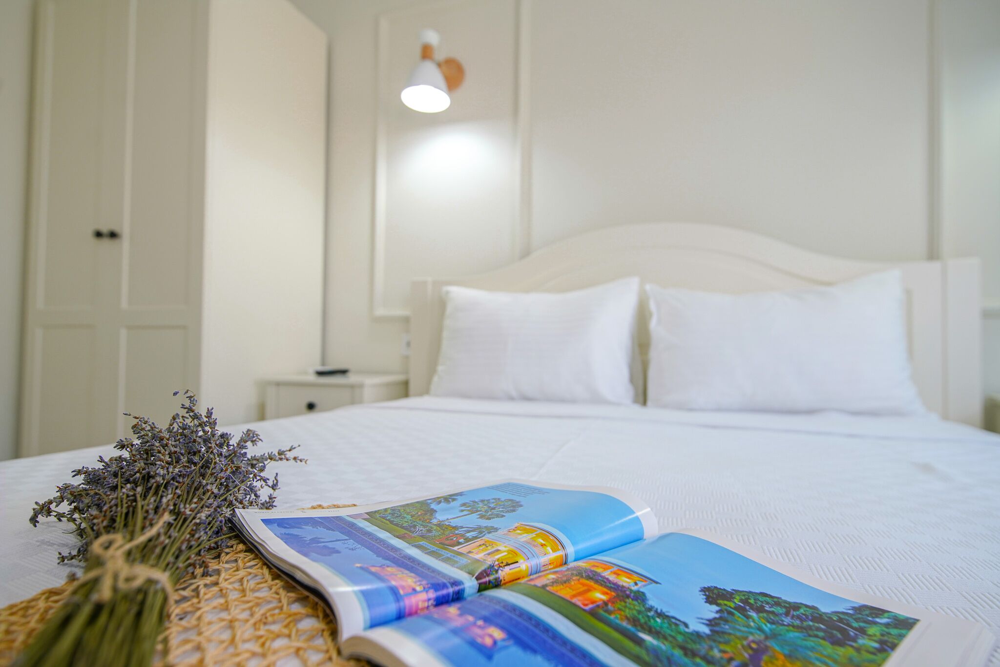 Un lit bien fait, avec des draps blancs, un livre et un bouquet de lavande posés sur un tapis en fibres naturelles.