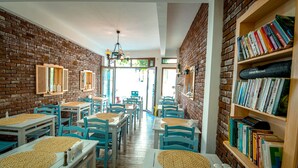 Restaurante com paredes de tijolo, estantes de madeira e cadeiras azuis. As mesas têm jogos americanos trançados.