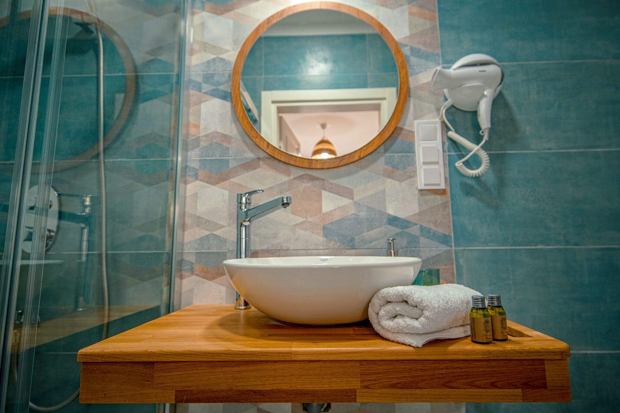 Un baño con tocador de madera, un lavabo blanco, un espejo redondo y un secador de pelo en la pared.