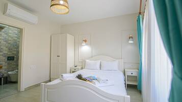 Et soveværelse med en hvid seng, et sengebord, et garderobeskab, en hængende lampe og et badeværelse, der kan anes gennem en åben dør.