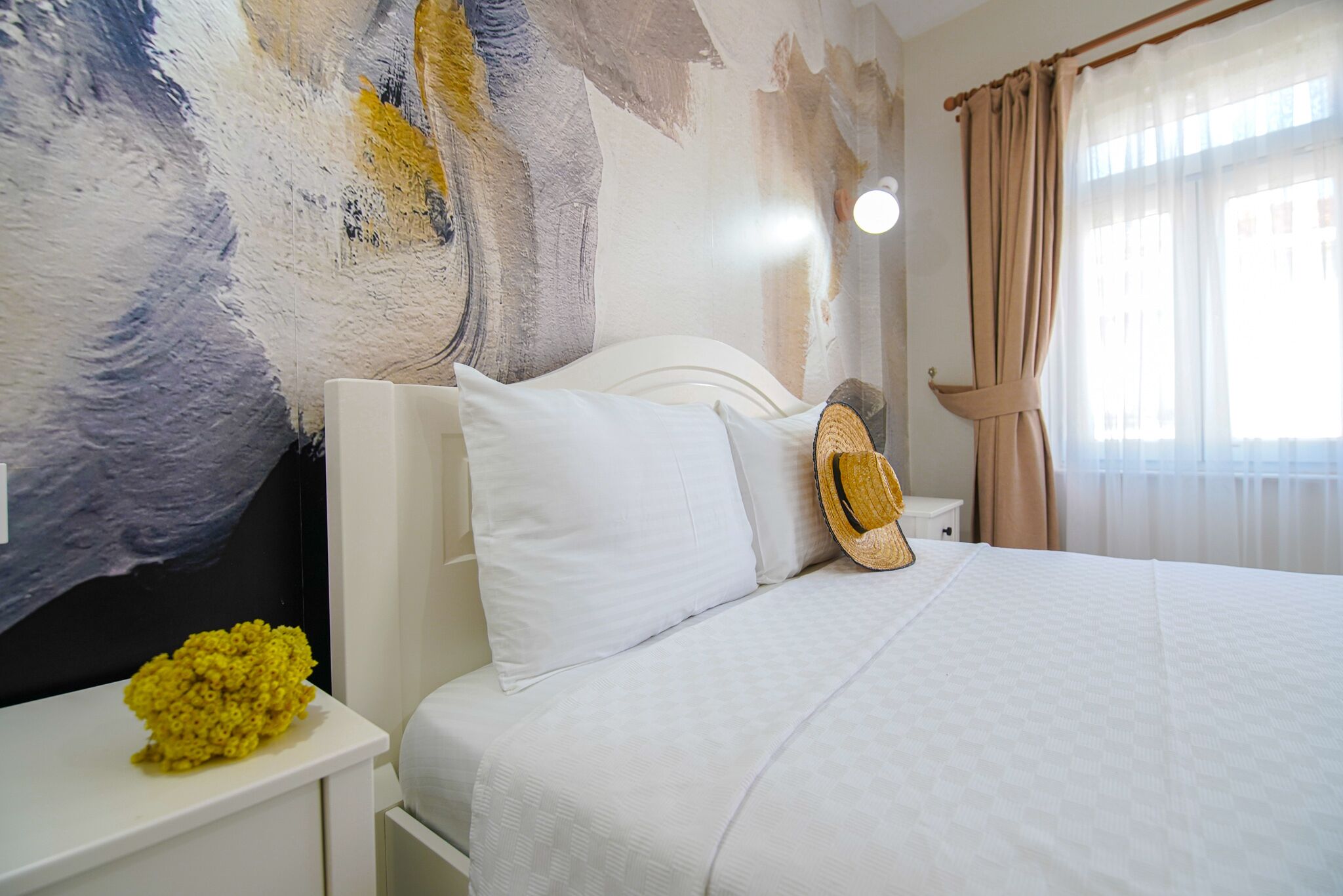 A hotel room with a bed, white nightstands, a wall mural, and a window with curtains.