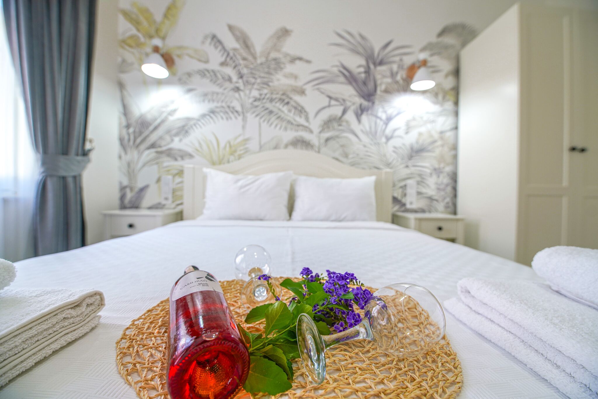 A neatly made bed with a woven placemat, a glass decanter, and a glass bottle of red liquid.
