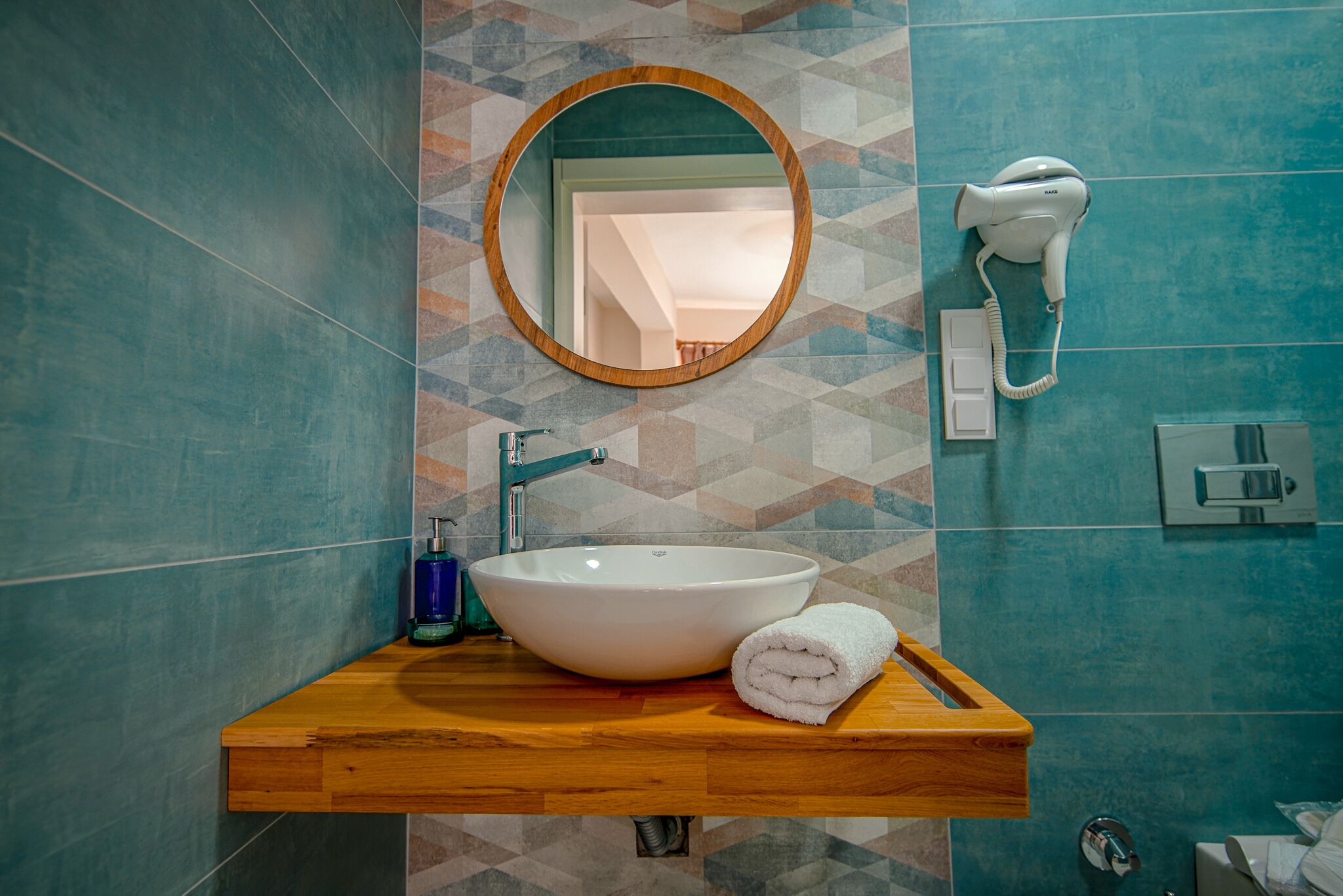 A bathroom with a wooden vanity, a white sink, a towel, a round mirror, a hairdryer, and a geometric-patterned wall.