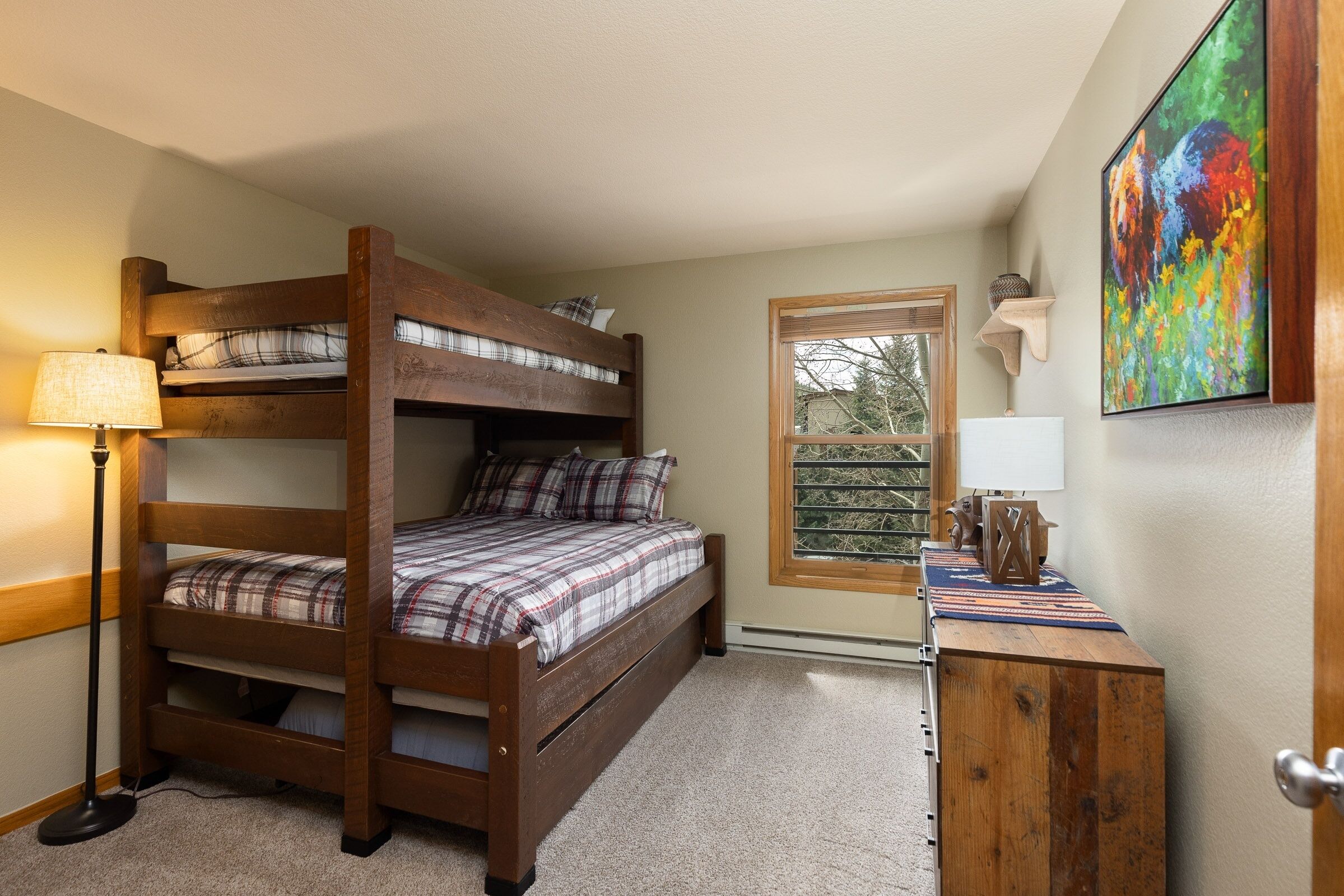 A bunk bed room with a window, a desk with a lamp, and a painting on the wall.