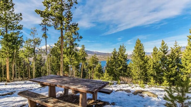 A wooden picnic table in a snowy forest with a clear view of mountains in the background.