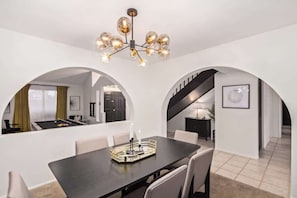 A dining area with a dark wooden table, white chairs, and a chandelier. There are arched doorways leading to other rooms. - Tiki Paradise w/Htd. Pool, Putt Grn. & Tiki Bar (Phoenix)