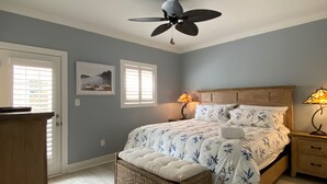 Un dormitorio con cama, dos lámparas de noche, ventilador de techo, banca de mimbre y una puerta con persianas.